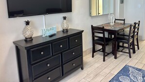Uma área de refeições com televisão na parede, um armário de madeira escura com gavetas, uma mesa com cadeiras e um espelho decorativo.