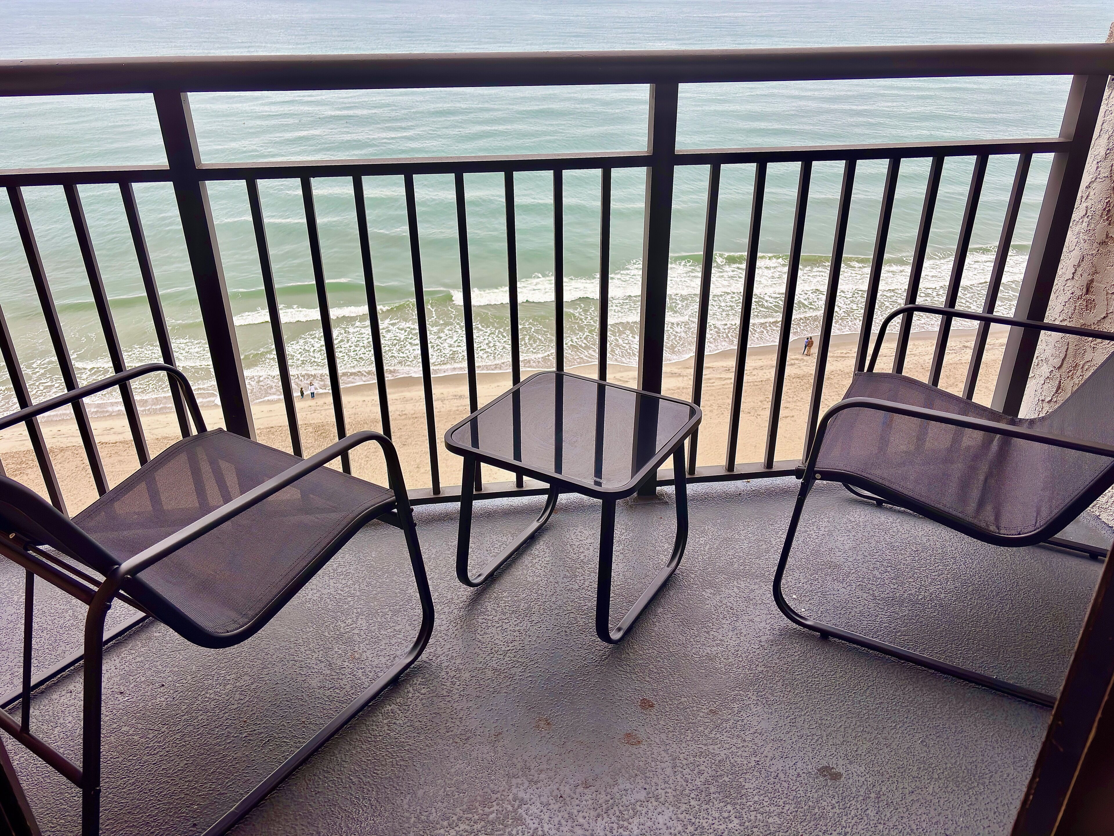 A balcony with two chairs and a small table, overlooking a beach with waves and people in the distance.