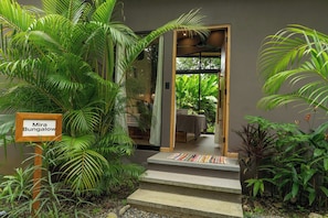 Un letrero de madera con la inscripción 'Mira Bungalow', un conjunto de escalones de concreto y abundante follaje verde.