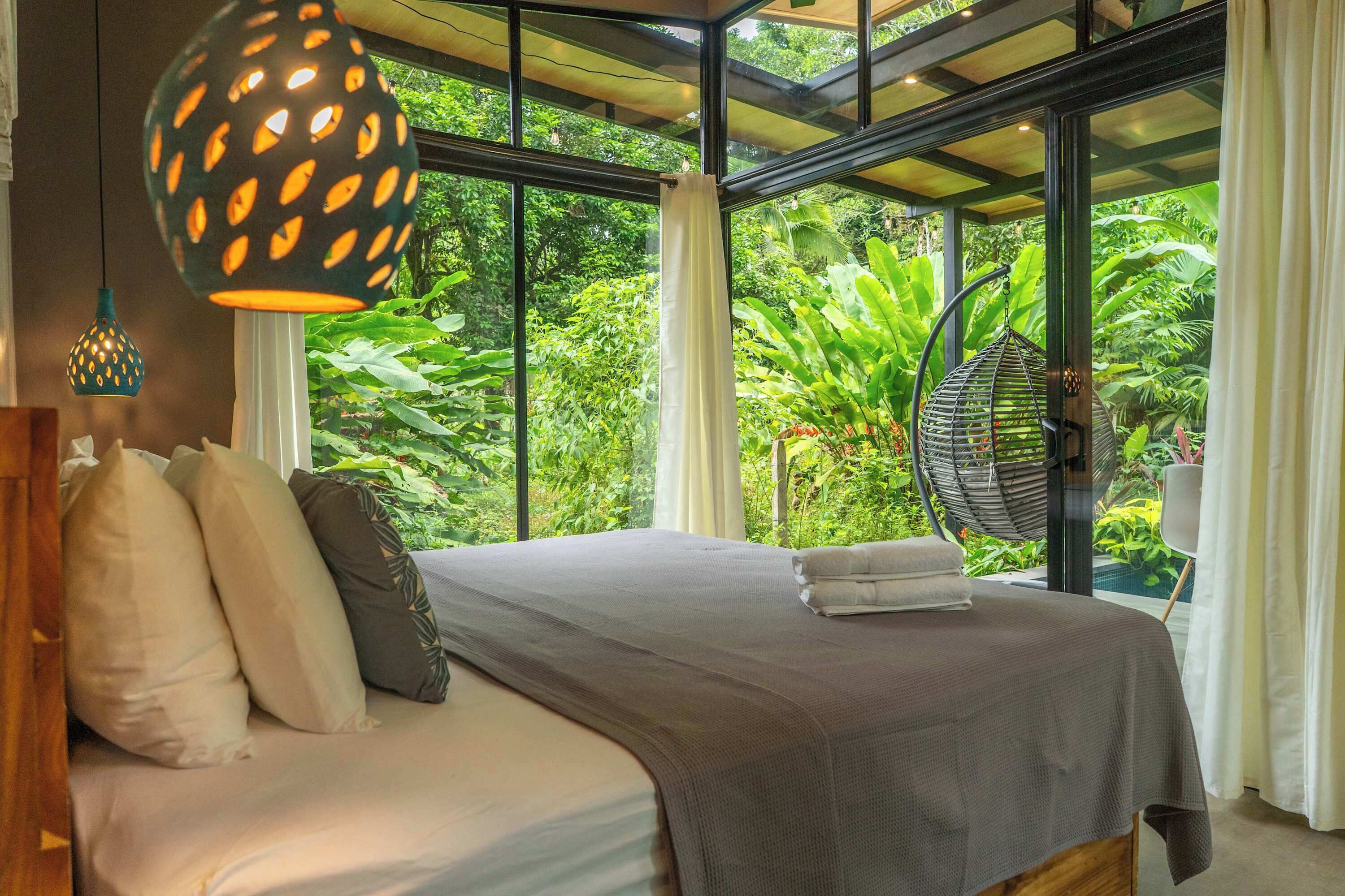 Un dormitorio con cama, almohadas, una lámpara colgante y vistas a abundante vegetación a través de amplios ventanales.