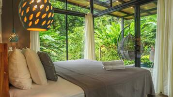 Un dormitorio con cama, almohadas, una lámpara colgante y vistas a abundante vegetación a través de amplios ventanales.