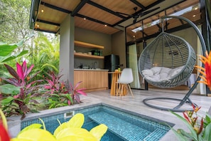 Una cocina exterior moderna con una silla colgante, piscina y abundante vegetación.