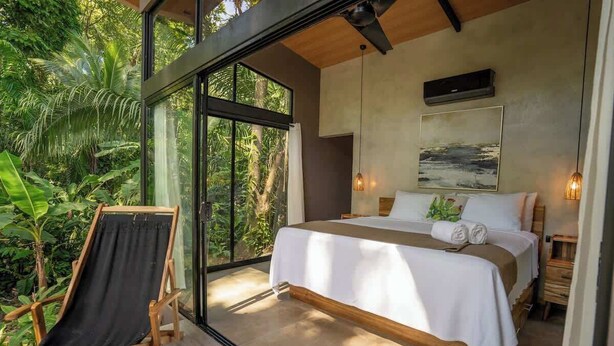 A bedroom with a large bed, a wooden chair, and a view of lush greenery through large windows. - Villa Asencia w/ pool & AC near the beach (Punta Uva)