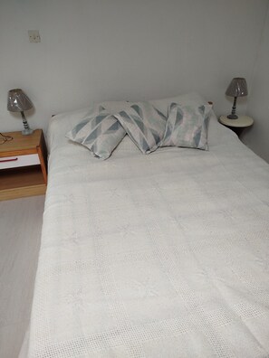 A neatly made bed with a white knitted blanket, two pillows with a geometric pattern, a bedside table with a lamp, and a wooden nightstand with a book and glasses. - Apartment (Argelès-sur-Mer)