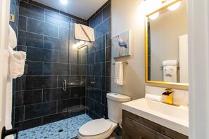 A modern bathroom with dark tiles, a glass-enclosed shower, a toilet, and a sink with a gold faucet. - Stylish King Suite Steps to Dining & Parks (San Diego)