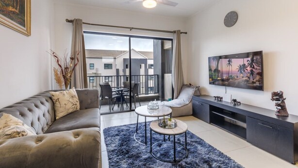 A modern living room with a sofa, a coffee table, a TV, and a balcony with a view of houses. - Haus of Salt (Ballito)