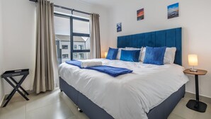 A modern hotel room with a large bed, a ceiling fan, a small table with a lamp, and a window with curtains. - Haus of Salt (Ballito)