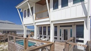 Een twee verdiepingen tellend huis aan het strand met een zwembad, ligbedden en een houten omheining.