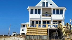 Een wit, twee verdiepingen hoog huis met een balkon, gelegen aan een zandstrand met een houten omheining en een trap die naar de ingang leidt.