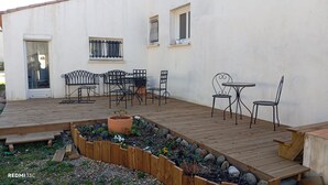 Una terraza de madera con una mesa y sillas de metal, rodeada de un jardín con diversas plantas y una maceta.