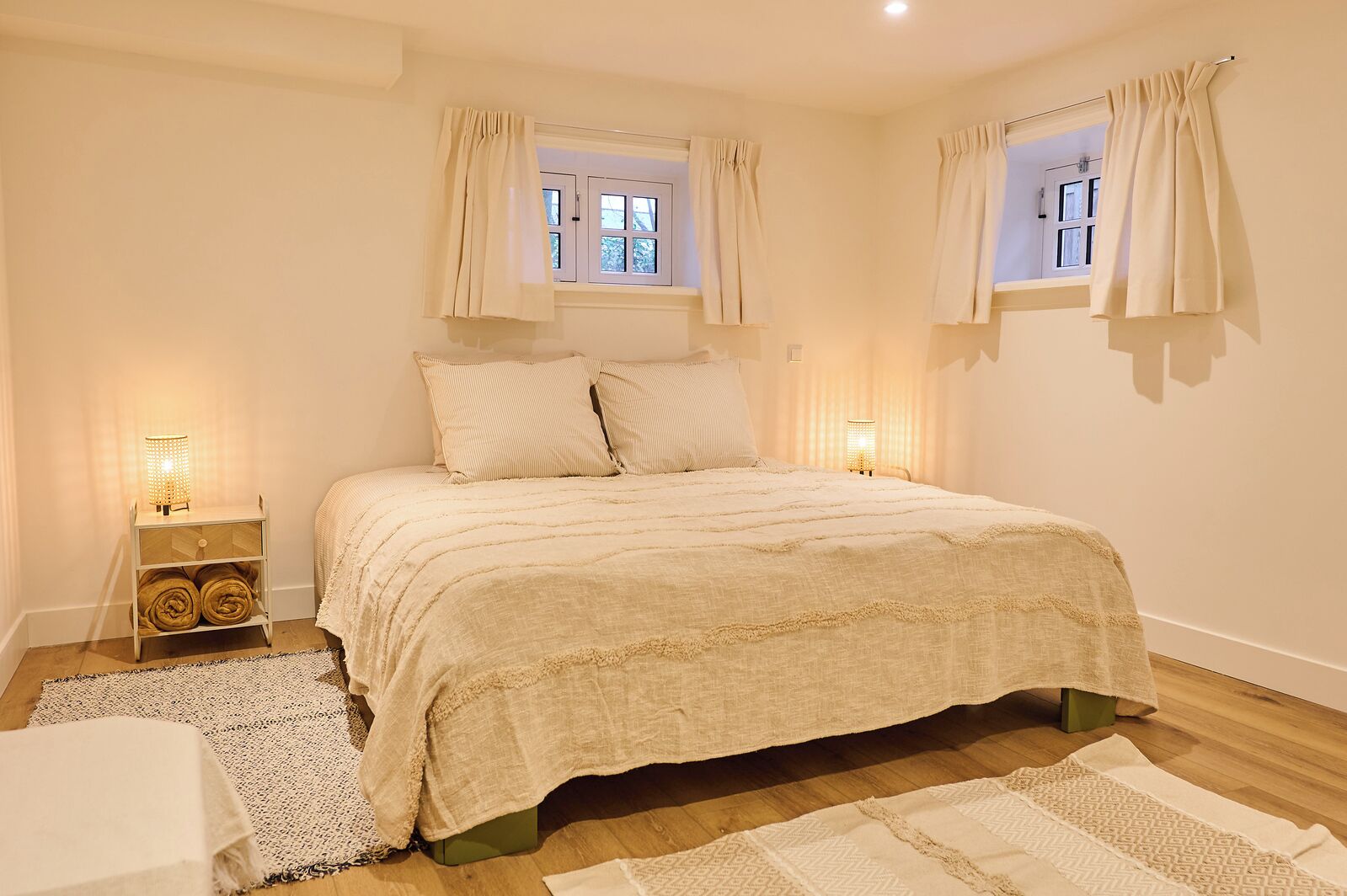 A bedroom with a large bed, two bedside lamps, a window with curtains, and a bedside table with a lamp.