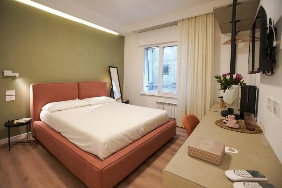 A hotel room with a bed, a desk with a chair, a window, and a radiator.