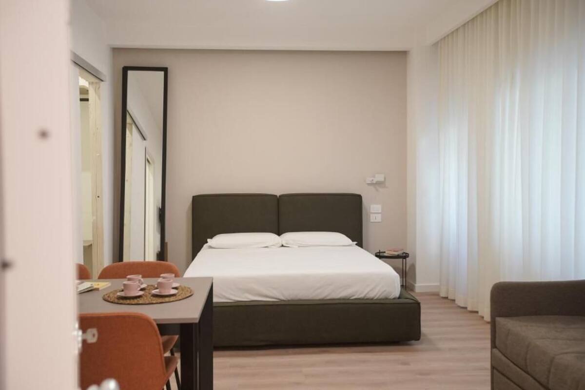 A modern hotel room with a bed, a small table with cups, a chair, and a sofa.