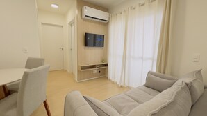 A modern living room with a sofa, a TV mounted on the wall, a dining table with chairs, and a kitchen area with a counter and cabinets. - Solarium Beach Club - Apartamento T1.406 (Itapoá)