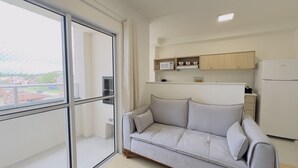 A modern apartment with a sofa, kitchenette, and a large window with a view of buildings. - Solarium Beach Club - Apartamento T1.406 (Itapoá)
