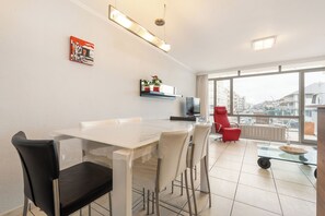Eine moderne Küche mit weißer Arbeitsplatte, einem schwarzen Stuhl und einem roten Stuhl. Ein Fenster bietet Blick auf Gebäude und es gibt einen Balkon.