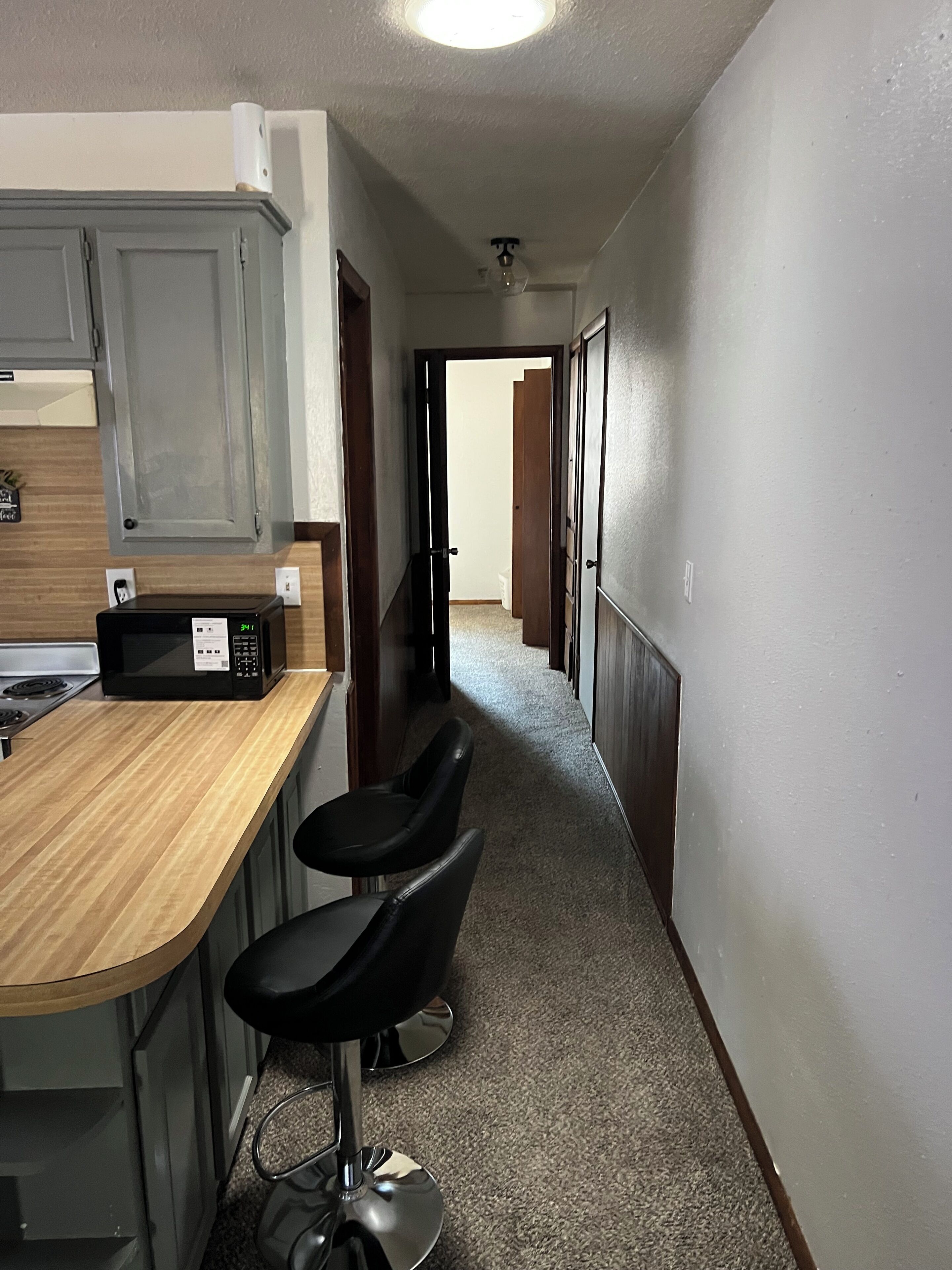 A narrow hallway with a kitchen area on one side, featuring a microwave, a wooden countertop, and two black bar stools.