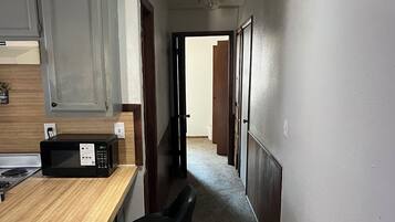 A narrow hallway with a kitchen area on one side, featuring a microwave, a wooden countertop, and two black bar stools.
