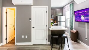 A modern kitchen with a built-in microwave, stove, and refrigerator. There is a small island with bar stools, a trash can, and a wall-mounted TV displaying a cityscape. - Chic Studio – Steps to Dining, Wi-Fi & A/C (San Diego)