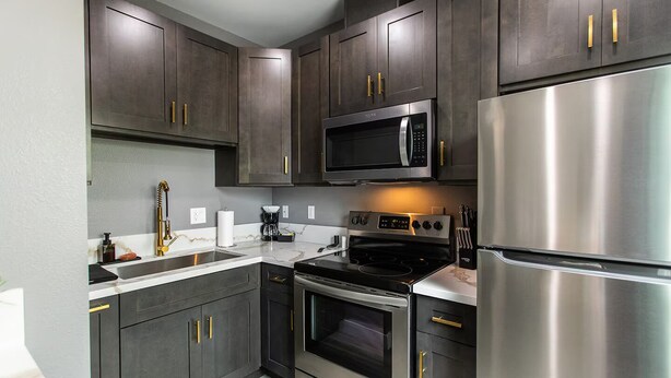 A modern kitchen with dark cabinets, stainless steel appliances, and gold handles. - Chic Studio – Steps to Dining, Wi-Fi & A/C (San Diego)