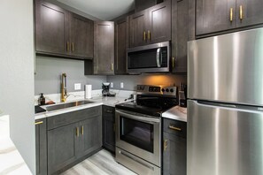 A modern kitchen with dark cabinets, stainless steel appliances, and gold handles. - Chic Studio – Steps to Dining, Wi-Fi & A/C (San Diego)
