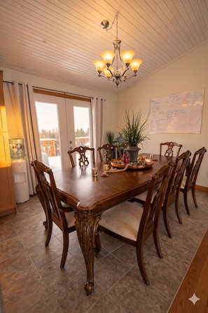 Une salle à manger avec une table en bois, six chaises, un lustre, une plante en pot et un tableau blanc accroché au mur.