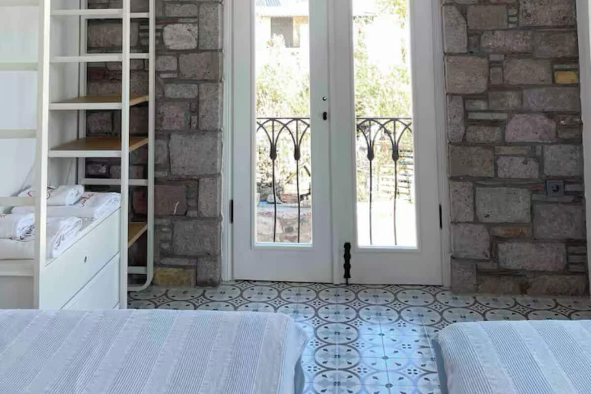 Une chambre avec une armoire blanche, un lit avec une literie à motifs, un luminaire suspendu en osier et un mur en pierre.