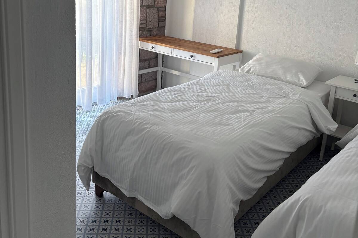 Une chambre à coucher comprenant un lit, des tables de chevet, un bureau en bois, un mur d’accent en pierre et un luminaire suspendu.