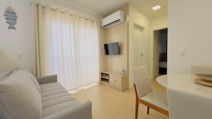 A modern living room with a sofa, a TV mounted on the wall, a dining table with chairs, and an air conditioning unit. - Solarium Beach Club - Apartamento T1.505 (Itapoá)