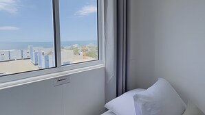 A hotel room with a large window, a bed with white bedding, and a view of the sea and buildings. - Solarium Beach Club - Apartamento T1.505 (Itapoá)