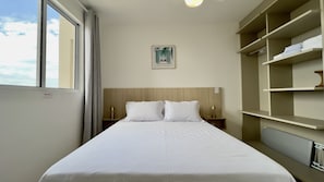 A hotel room with a bed, a nightstand, a lamp, a shelf with folded towels, and a window with curtains. - Solarium Beach Club - Apartamento T1.505 (Itapoá)