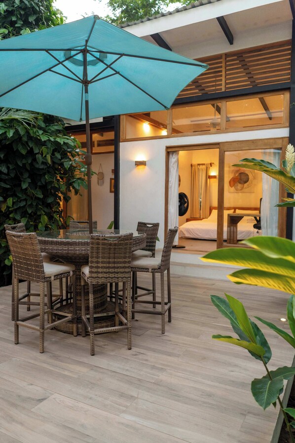 Un coin repas avec des meubles en osier et un grand parasol, une chambre avec un lit et une table de chevet, et une vue sur un bâtiment aux poutres en bois et doté d’un balcon.