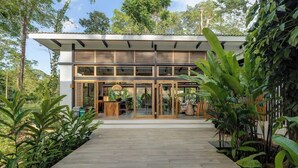 Un edificio moderno con terraza de madera, puertas de cristal y abundante vegetación a su alrededor.