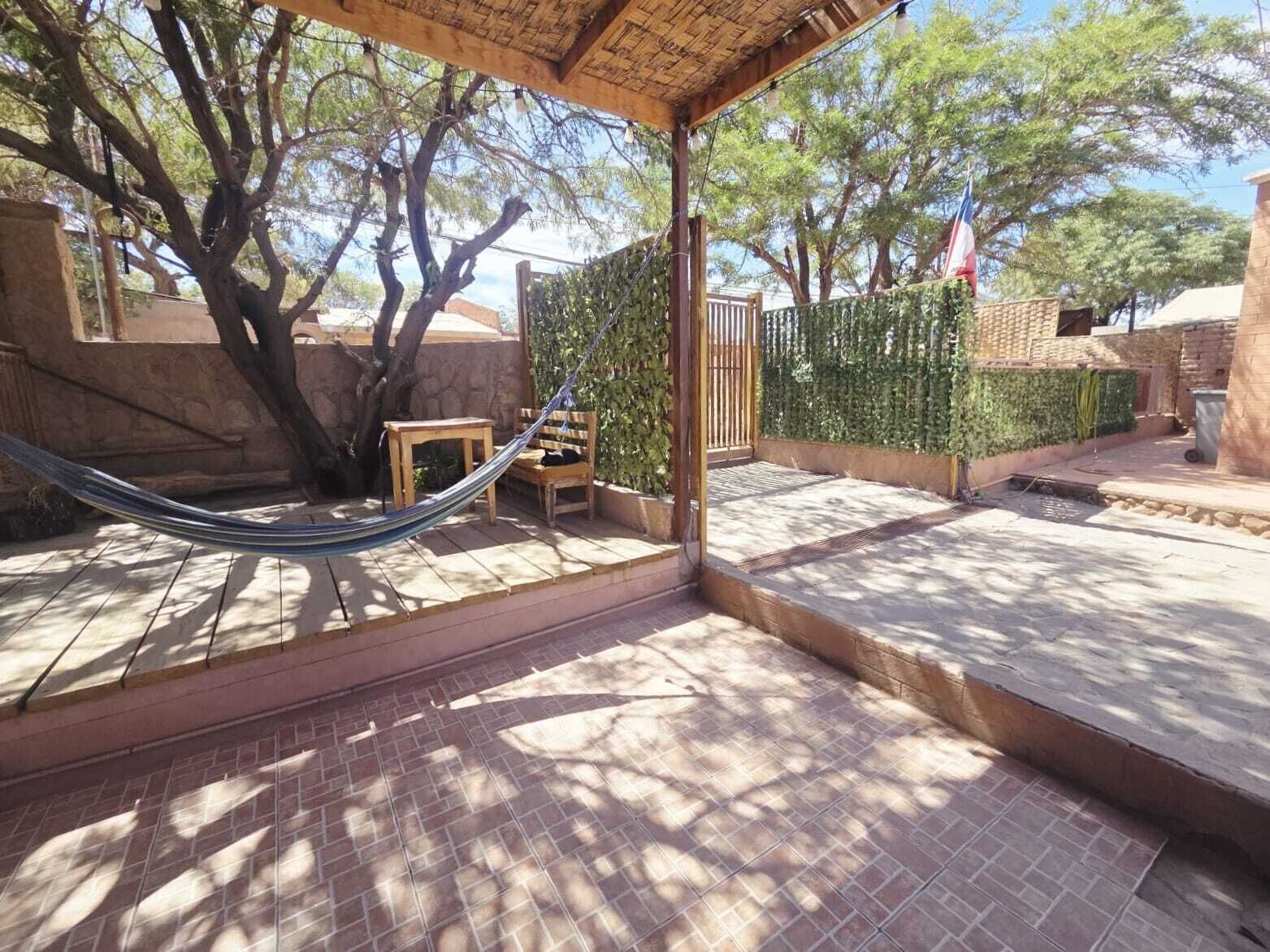 Un patio cubierto con una hamaca, una mesa de madera y sillas, rodeado de árboles y un muro de piedra.