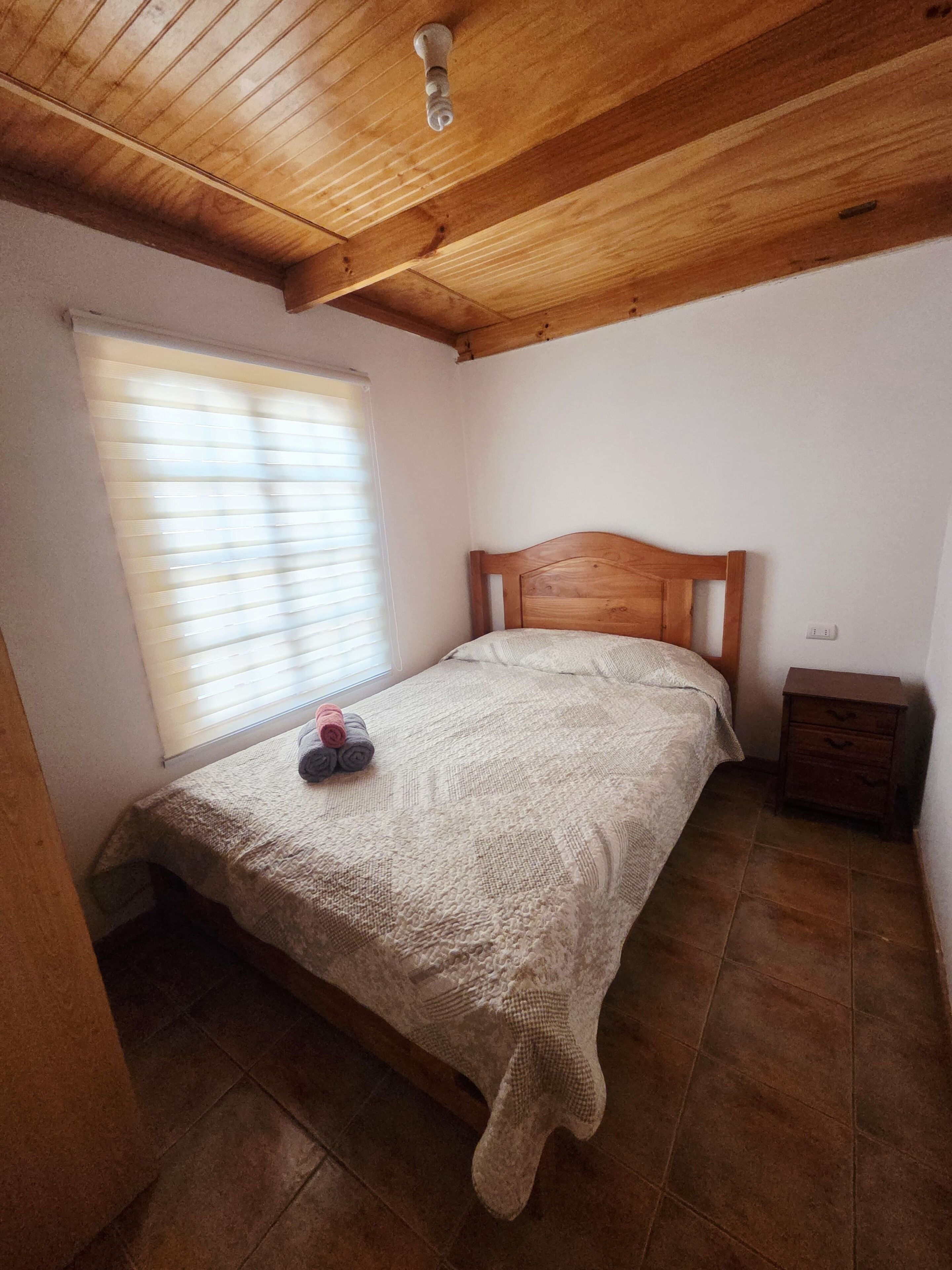A bedroom with a wooden ceiling, a bed with a knitted blanket, a nightstand, and a window with blinds.