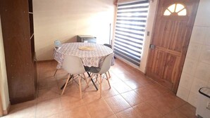 Un comedor con una mesa redonda, sillas, mantel estampado, una ventana con persianas y una puerta de madera.