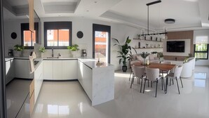 A modern kitchen with a central island, a dining area with a wooden table and chairs, and a living space with a TV and plants. - 3 Bed Villa - Sleeps 6 - Private Pool - Parking (Rojales)