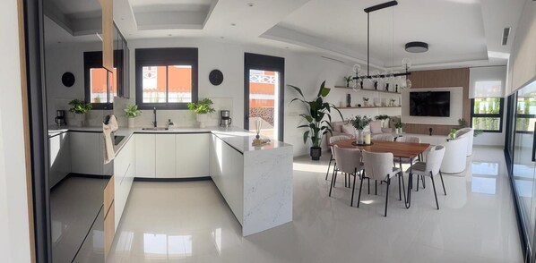 A modern kitchen with a central island, a dining area with a wooden table and chairs, and a living space with a TV and plants. - 3 Bed Villa - Sleeps 6 - Private Pool - Parking (Rojales)