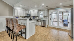 A modern kitchen with white cabinetry, a central island, and a dining area with woven chairs. - Walk to Beach & Parks! Coastal Haven in Oak Island (Oak Island)