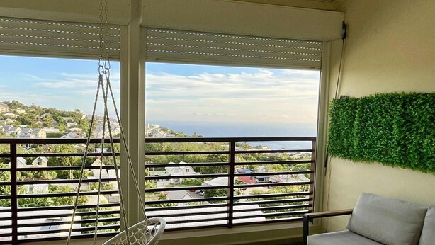 Ein Balkon mit einer Hängematte, einer Couch und einem Tisch, mit Blick auf eine Wohngegend mit Häusern und Grünflächen.