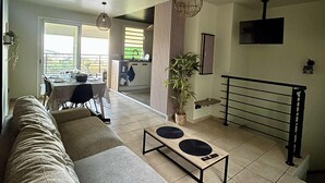 Ein modernes Wohnzimmer mit einer Couch, einem Couchtisch und einem Essbereich mit Tisch und Stühlen. Eine Treppe mit Geländer, ein Blumentopf und eine Pendelleuchte sind ebenfalls vorhanden.