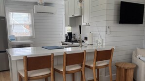 Une cuisine moderne avec des meubles blancs, un îlot central doté de tabourets, un téléviseur à écran plat fixé au mur et une fenêtre avec des stores.