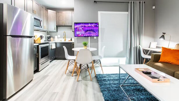 A modern kitchen with stainless steel appliances, a dining area with white chairs, a small round table, and a TV mounted on the wall. - Spacious Studio – Balcony, Desk, A/C & Kitchen (San Diego)