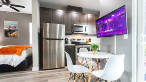 A compact kitchen with a stainless steel refrigerator, microwave, and stove, a dining area with a white table and chairs, and a TV mounted on the wall displaying a cityscape. - Spacious Studio – Balcony, Desk, A/C & Kitchen (San Diego)