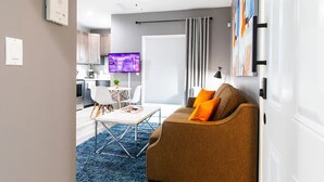 A modern living room with a sofa, a coffee table, and a TV. There is a kitchen in the background with cabinets and a window. - Spacious Studio – Balcony, Desk, A/C & Kitchen (San Diego)