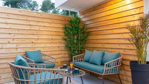 Een overdekt terras met houten lambrisering, rieten stoelen en een tafel, omringd door plantenpotten.