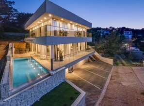 A modern two-story house with a glass exterior, a swimming pool, and a stone retaining wall.