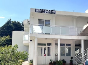 Ein modernes Gebäude mit einem Schild, auf dem "APARTMANI" und Kontaktdaten stehen. Es hat einen Balkon mit Metallgeländern und Blumentöpfen.