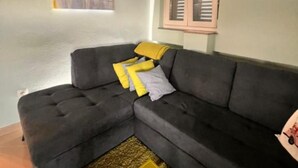 Ein graues Ecksofa mit gelb-weißen Kissen, ein kleiner gelber Hocker und ein gemustertes Teppich.