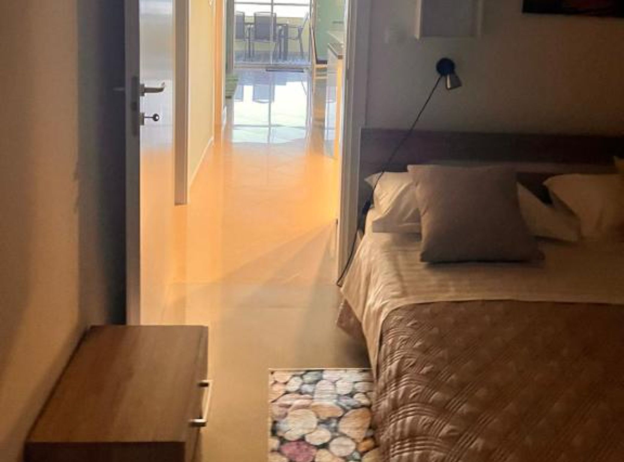 Une chambre à coucher comprenant un lit, une table de chevet, une lampe et donnant sur la salle à manger par une porte ouverte.